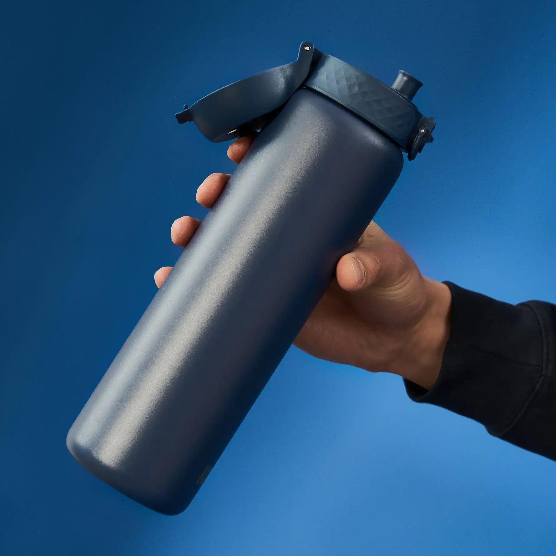 A dark-blue water bottle is being held, its flip-top open. The hand and a portion of a dark sleeve are visible against a solid blue background.