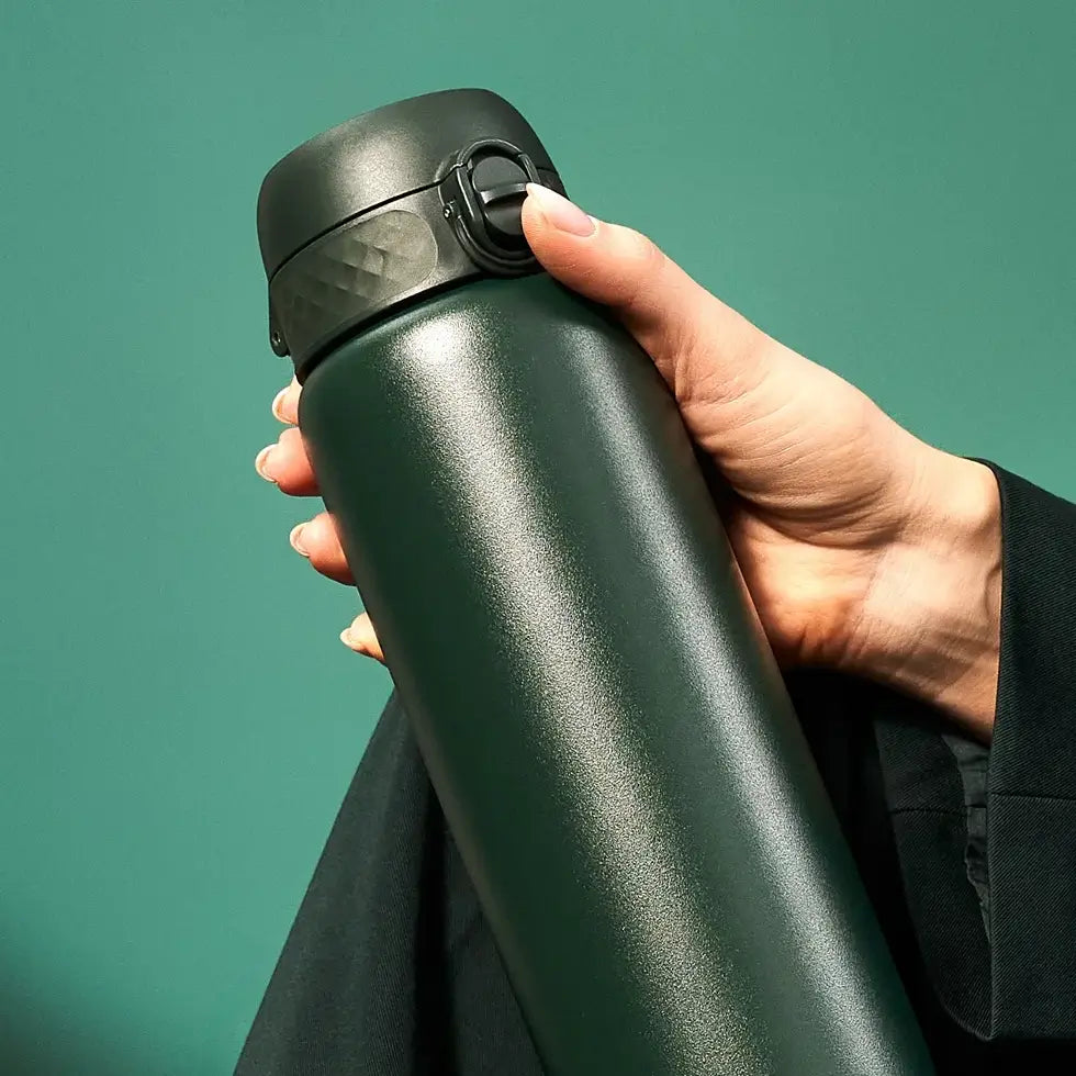 A dark-green thermos is held in a hand against a teal background.